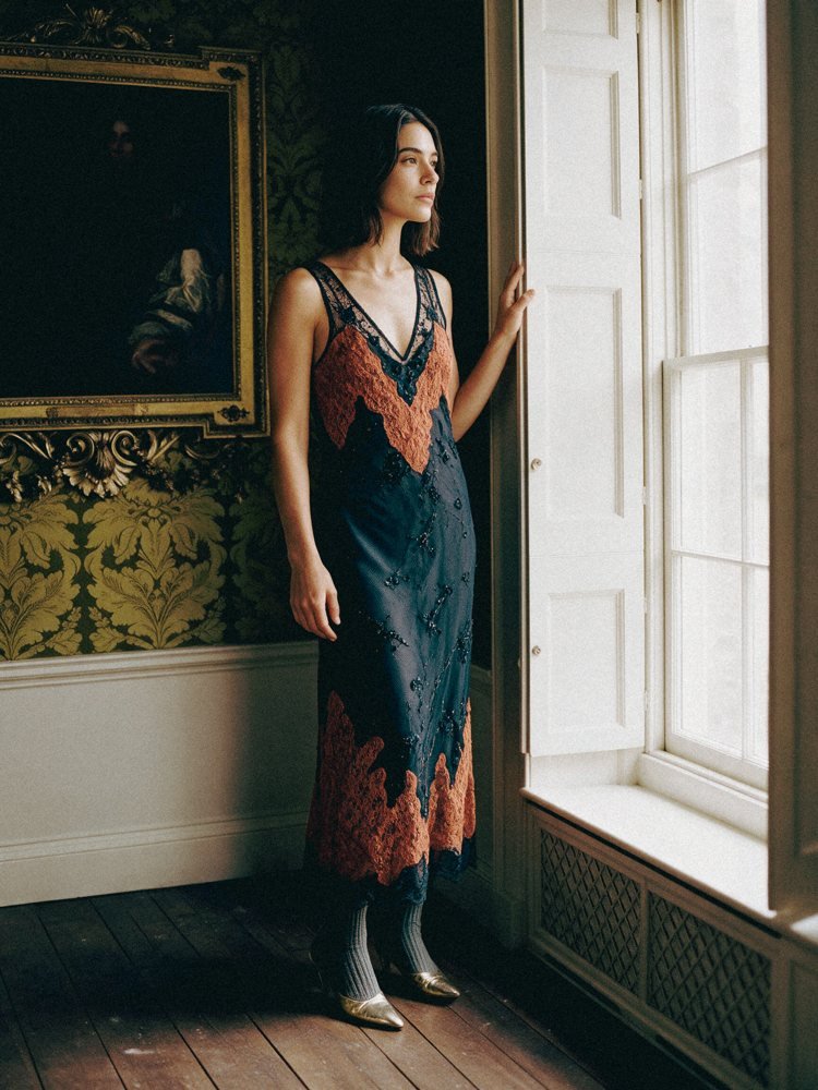 Joana com o mesmo vestido de pé junto à janela de uma sala antiga